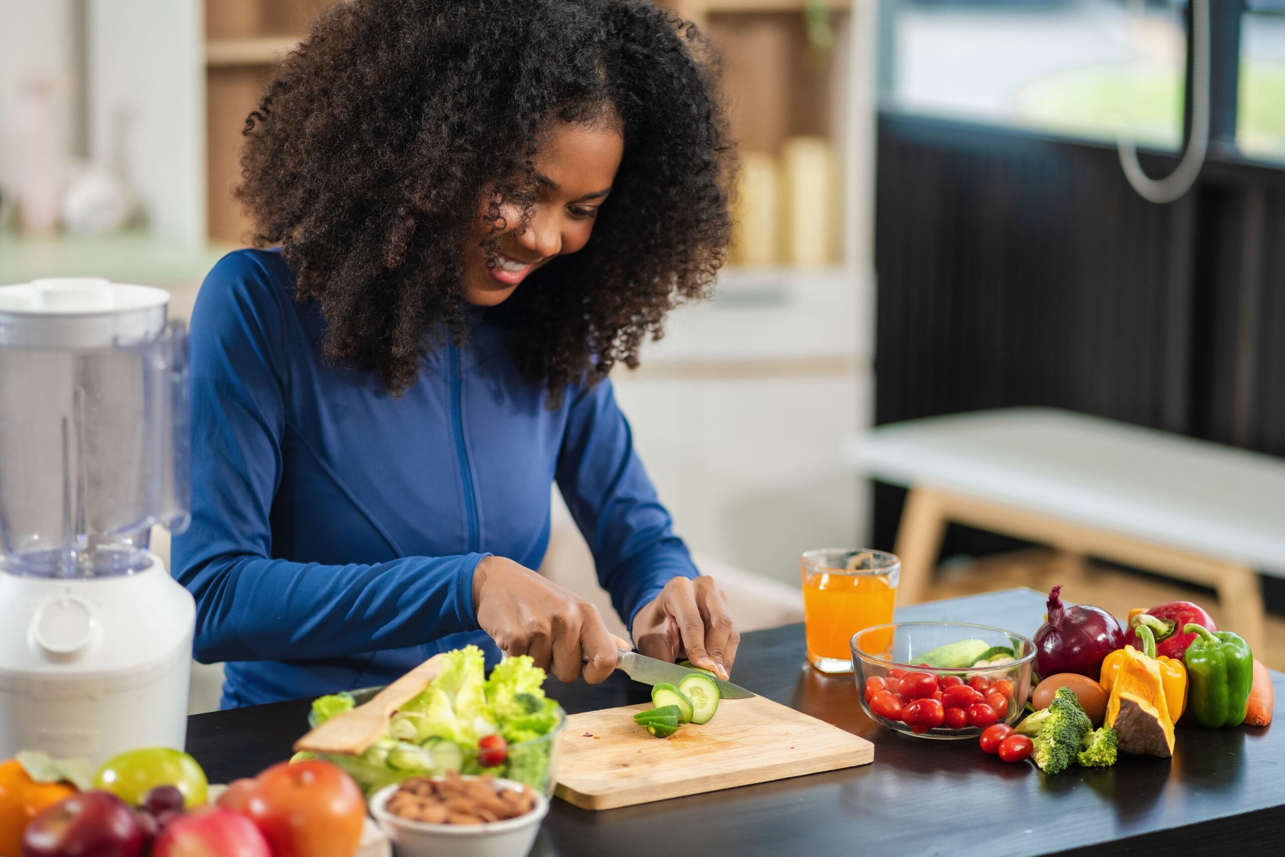 Black woman standing at the counter cutting a cucumber with other vegetables laid out on the counter top, practicing healthy living